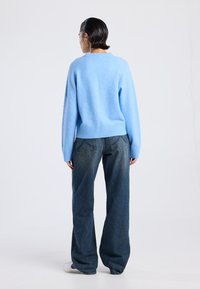 Pull en tricot bleu ciel avec ourlet et poignets côtelés, associé à un jean large bleu foncé. Texture douce et coupe décontractée, vu de derrière.