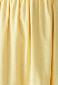 Keltainen kangas hienolla verkkokudoksella, roikkumassa pehmeillä pystysuorilla poimuilla ja lempeillä varjoilla.