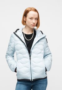 Νεαρός με κοντά κόκκινα μαλλιά, που φοράει ανοιχτό μπλε μπουφάν με καπιτονέ λεπτομέρειες πάνω από μαύρο πουκάμισο και μαργαριταρένιο κολιέ, με τα χέρια στις τσέπες, μπροστά σε λευκό φόντο.