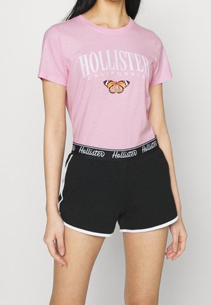 Kobieta w różowym t-shircie Hollister z motylem i czarnych szortach Hollister z białym obszyciem i paskiem z logo.