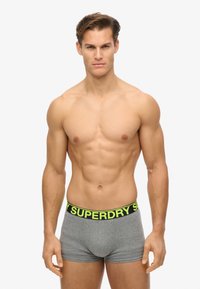 Szare bokserki z czarnym i neonowozielonym paskiem "SUPERDRY", charakteryzujące się dopasowanym krojem i miękką teksturą. Model ma umięśnioną sylwetkę.