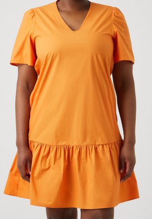 Kobieta ubrana w pomarańczową sukienkę z krótkim rękawem, dekoltem w serek i marszczonym falbanowym dołem, stojąca na tle jasnej, gładkiej ściany.