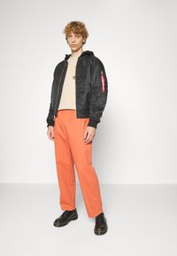 Zwarte bombere jacket met capuchon, ritssluiting aan de voorkant en rode accenten op de mouw; beige T-shirt, oranje broek en zwarte schoenen.