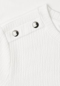 Gros plan sur un tissu blanc côtelé avec un tricot texturé et deux boutons argentés brillants près de l'encolure arrondie sur l'épaule d'un vêtement.