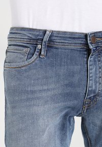Hellblaue Denim-Jeans mit tailliertem Schnitt, Fünf-Taschen-Design und sichtbaren Nahtakzenten. Die Textur ist glatt mit einem leicht abgenutzten Aussehen.