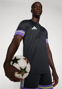 Zwart voetbalshirt met paarse en groene gestreepte mouwen, Adidas-logo op de borst. Model houdt een wit en zwart voetbal met sterpatroon vast.