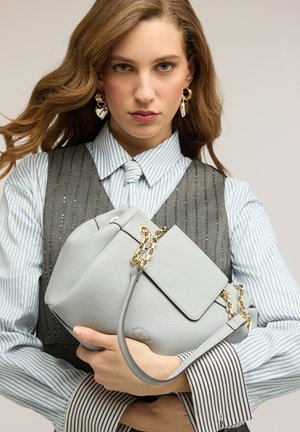 Ung kvinde med bølget brunt hår holder en lys grå læderhåndtaske med guldkædedetaljer, iført stribet skjorte og mørkegrå vest.