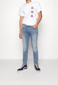 Vit t-shirt med färgglad text och grafik, kombinerad med ljusblå jeans och svarta högskaftade sneakers med vita sulor.