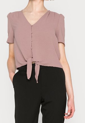 Kvinde iført lyserød kortærmet knappet bluse bundet i taljen og sorte bukser med høj talje, stående med den ene hånd i lommen.