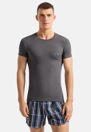 Bărbat tânăr purtând un tricou strâmt gri închis cu mânecă scurtă și un mic logo și pantaloni scurți albaștri în carouri, stând în fața unui fundal simplu.