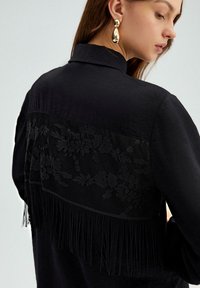 Zwarte shirt met een getextureerd bloemenkantpaneel op de rug, versierd met franjes langs de zoom. Gladde stofstructuur en kraag.