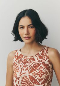 Ærmeløs bluse i cremefarvet stof med røde blomster mønstre. Har en rund halsudskæring og en løs pasform, der fremhæver et struktureret design.