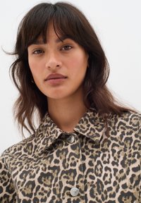 Jeansjacka med leopardmönster som har en kragliknande hals, silverfärgade knappstängningar, samt ett texturerat tyg i en ljusbeige och brun färgsättning.