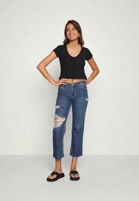 Mulher de pé com as mãos nos quadris, vestindo uma t-shirt curta preta, jeans rasgados azuis, sandálias pretas, sorrindo contra um fundo simples.