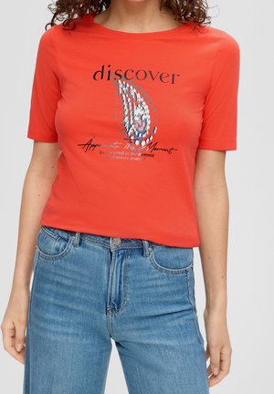 Vrouw draagt een rood T-shirt met korte mouwen met abstract ontwerp en tekst "discover", gecombineerd met blauwe jeans met hoge taille.