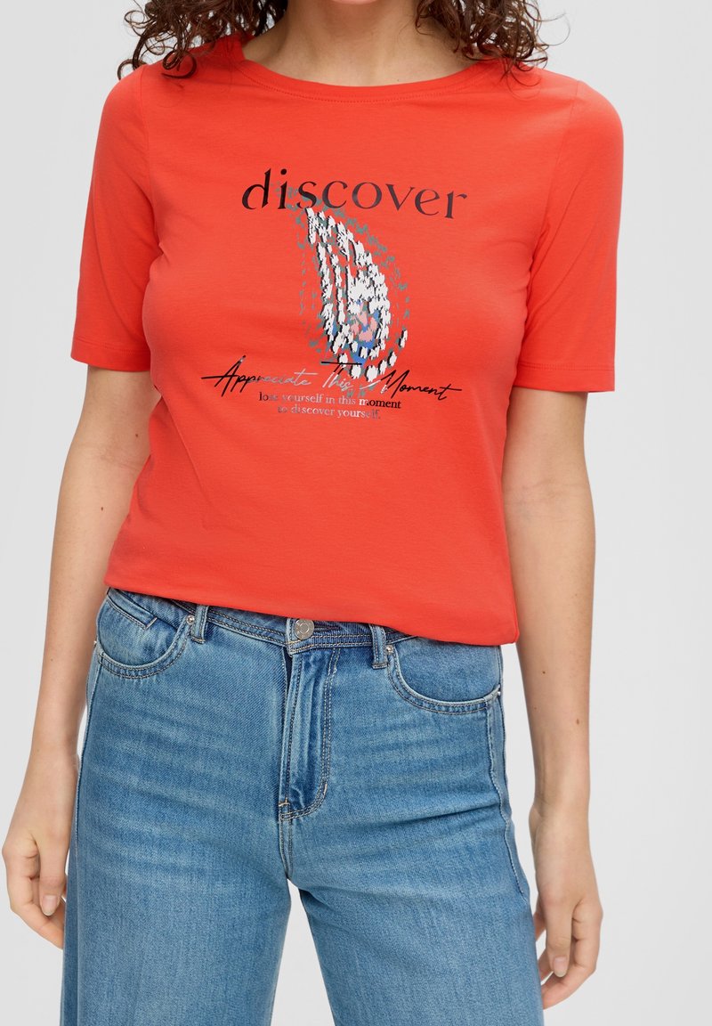 Femme portant un t-shirt rouge à manches courtes avec un design abstrait et le texte "discover", associé à un jean bleu taille haute.