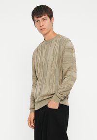 Beige striktrøje med strukturerede striber, rund halsudskæring og ribbede manchetter. I kombination med sorte bukser står modellen foran en hvid baggrund.