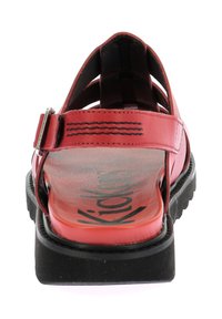 Sandal de material sintético rojo con diseño abierto, correa ajustable en el tobillo y suela de goma negra. Presenta el destacado logo "Kickers" en el interior.