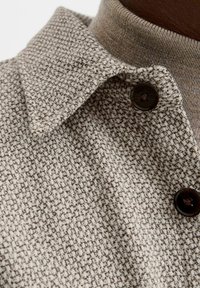 Skjorte med krave i et tekstureret beige stof, der har et subtilt mønster og mørkebrune knapper mod en lysere baggrund.