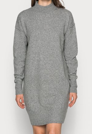 Kvinde iført en langærmet, grå strikket sweatervedkjole med ribbede manchetter og kant, stående mod en ensfarvet baggrund.