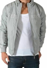 Lysegrå bomberjakke med lynlås foran, ribstrikkede ærmeafslutninger og "REDSKINS" broderet på brystet. Stoffet fremstår glat med et strømlinet design.