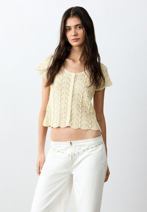 Young woman with long dark hair wearing a beige knitted short-sleeve top and white pants standing against plain background.
