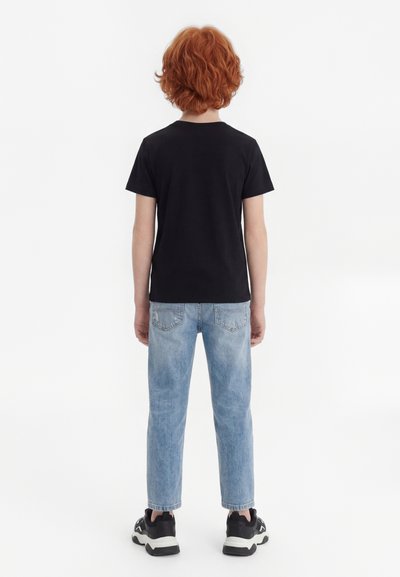 Copil cu păr roșcat și creț, văzut din spate, purtând un tricou negru, blugi albastru deschis și adidași negri pe un fundal alb.