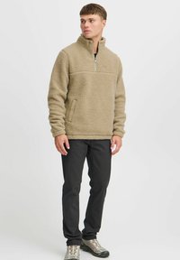 Beiger Fleece-Pullover mit Reißverschlusskragen, strukturiertem Stoff und einer Brusttasche; kombiniert mit schwarzen Hosen und grauen Schuhen.