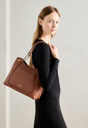 Carteira de mão castanha em pele com um acabamento texturizado, apresentando um bolso frontal, uma pega decorada com corrente e o logótipo na parte da frente. O modelo está a usar uma blusa preta.
