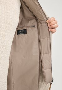 Gerecyclede wollen jas met een crèmekleurige textuur aan de buitenkant, een tan binnenvoering met een rits, en een label met merk- en materiaaldetails.