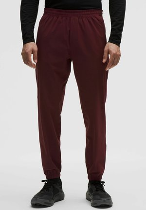 Vyras dėvi tamsiai raudonas elastingas laisvalaikio kelnes su kantu ties kulkšnimis, juodus sportinius batelius ir juodą ilgomis rankovėmis marškinėlį, stovintis tiesiai.