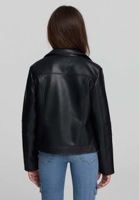 Persona con capelli lunghi e castani, che indossa una giacca di pelle nera e jeans di denim blu, girata di spalle su uno sfondo grigio uniforme.