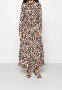 Donna che indossa un vestito floreale a maniche lunghe e lunghezza caviglia, con fiori rossi, bianchi e verdi, abbinato a sandali neri con tacco alto e cinturini.