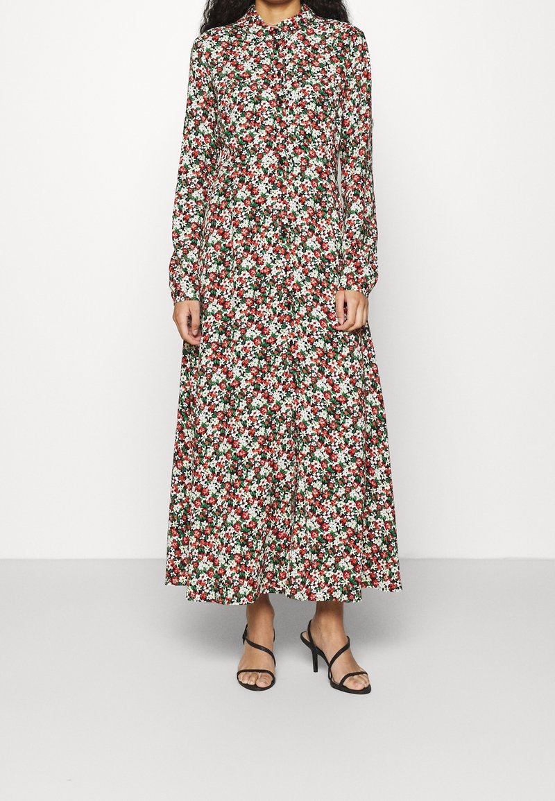 Donna che indossa un vestito floreale a maniche lunghe e lunghezza caviglia, con fiori rossi, bianchi e verdi, abbinato a sandali neri con tacco alto e cinturini.