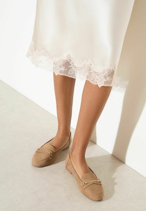 Kobieta nosząca beżowe zamszowe mokasyny ze złotymi elementami, w połączeniu z białą sukienką z misterną koronkową lamówką na dole.