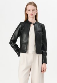 Giacca in pelle nera con colletto rotondo, zip frontale e taschino sul petto; indossata sopra un top in maglia chiara e pantaloni beige.