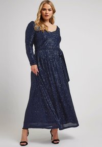 Abito lungo navy con paillettes, maniche lunghe, scollatura a barca e cintura legata in vita; scende fino al pavimento, con una texture di paillettes riflettenti.