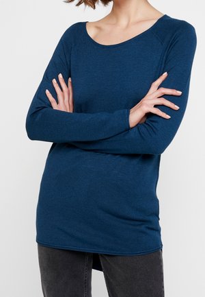 Persoon met een donkerblauw shirt met lange mouwen en donkergrijze broek, met de armen gekruist op de borst, tegen een effen achtergrond.