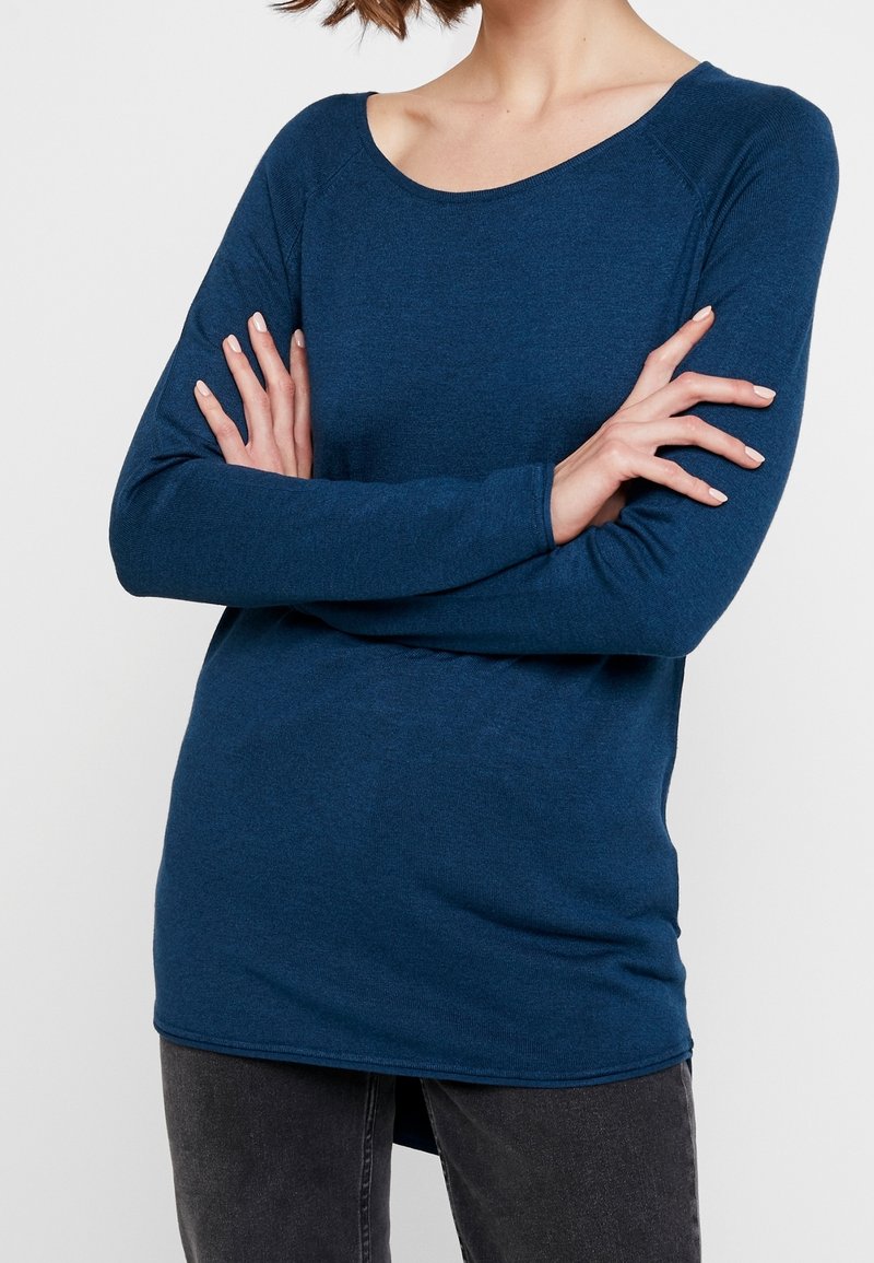 Personne portant un haut à manches longues bleu foncé et un pantalon gris foncé, les bras croisés sur la poitrine, devant un fond uni.