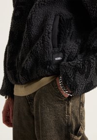 Schwarze Fleecejacke mit einer strukturierten, flauschigen Oberfläche und seitlichen Taschen. Getragen über einem beigen Hemd, kombiniert mit strukturierten dunklen Hosen und einem Kettenarmband.