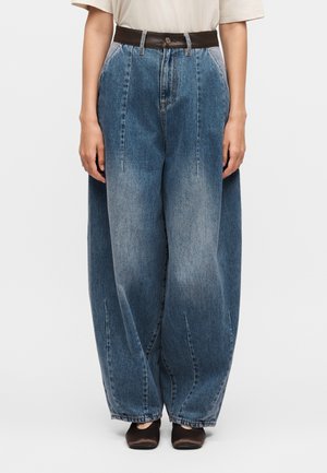 Jean en denim bleu délavé à jambes larges, taille haute et ceinture sombre, porté avec un haut blanc à manches courtes et des chaussures noires à bout carré.