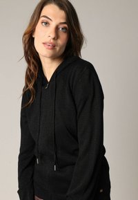 Zwarte gebreide hoodie met een trekkoord, voorzien van een gestructureerde afwerking en zilveren hardware accenten. De pasvorm is relaxed met lange mouwen.