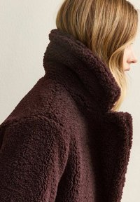 Profilo di una persona con capelli castano chiaro che indossa una giacca in pile testurizzato a collo alto, marrone scuro, su uno sfondo semplice.