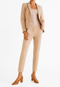 Beżowy, dopasowany żakiet i spodnie z gładkiego materiału, połączone z brązowymi butami na blokowym obcasie. Minimalistyczny design, prosty krój i neutralne kolory.