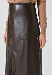 Brauner Leder-Maxi-Rock mit eng anliegendem Bund, Seitennähten und einem kleinen Metall-Logo-Akzent an der unteren Vorderseite. Glatte Textur.