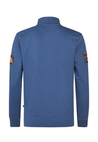 Felpa blu con maniche lunghe, colletto alto e polsini a coste. Presenta due toppe marroni sulle spalle. Tessuto morbido e strutturato.