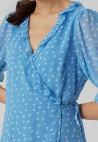 Blå omlotklänning med vitt blommönster, volangkrage och korta puffärmar. Har en knytning i midjan. Tillverkad av lätt, genomskinlig tyg.