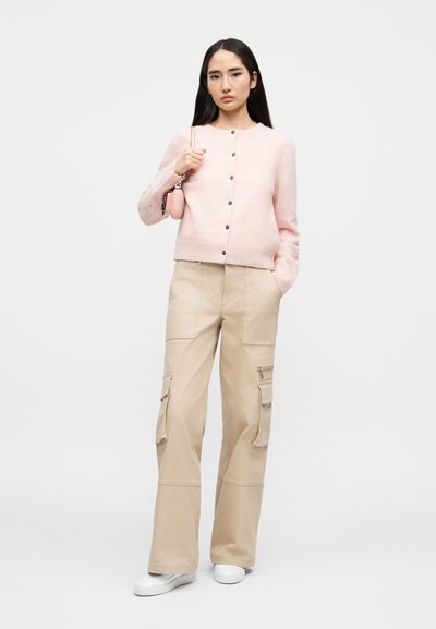Γυναίκα που φοράει ανοιχτό ροζ ζακέτα με κουμπιά, μπεζ παντελόνι cargo, λευκά αθλητικά παπούτσια και κρατά μια μικρή ροζ τσάντα στον ώμο της.