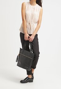 Donna che indossa un top beige senza maniche e pantaloni neri, mentre tiene in mano una grande borsa tote in pelle nera con cerniera e nappina.