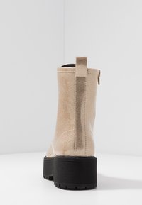 Glinsterende beige enkellaars met een gestructureerde afwerking, voorzien van een zwarte plateauzool, een ritssluiting aan de zijkant en een pull-tab aan de achterkant voor gemakkelijk aan- en uittrekken.